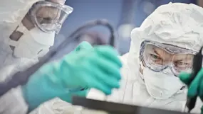Two researchers in white protective suits, masks, and goggles concentrate as they adjust lab equipment in a cleanroom.