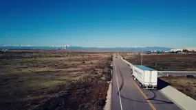 White semi‑truck with a long trailer driving on a quiet two‑lane road beside open fields, with distant mountains and a clear blue sky