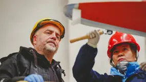 Two workers in hard hats and gloves look up as they work on the underside of a large red structure indoors; one holds a roller‑style tool while the other observes