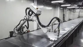 Industrial robotic arm with a tool head working along a long metal panel in a clean, fluorescent‑lit manufacturing cell